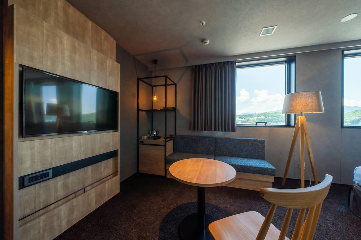 A modern hotel room with a large flat-screen TV, a round wooden table, a wooden chair, a sofa, and a window with a view of the outdoors.