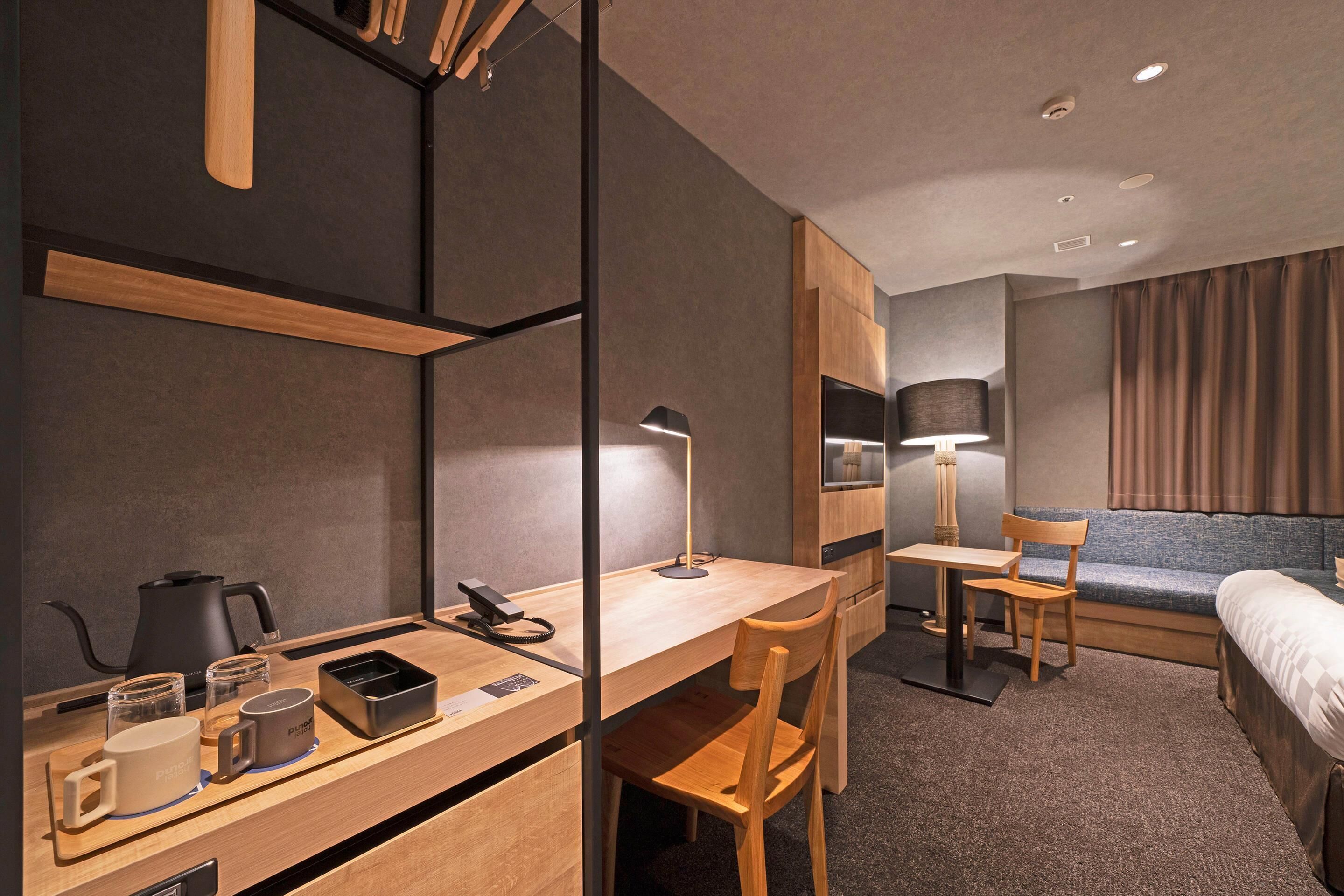 A modern hotel room with a wooden desk, a chair, a bed, a lamp, and a kettle.
