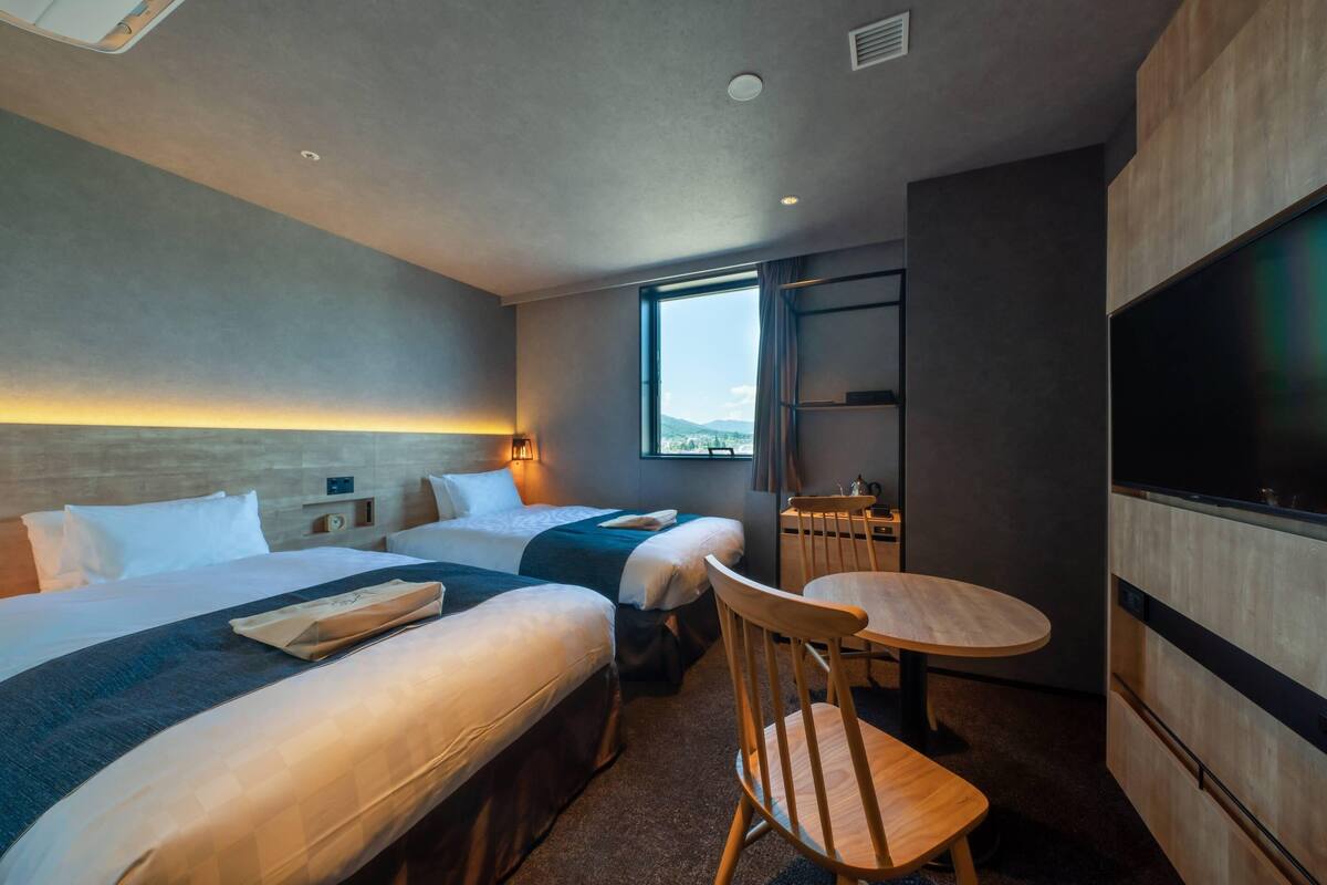 A hotel room with two beds, a dining table, a TV, and a window with a view.