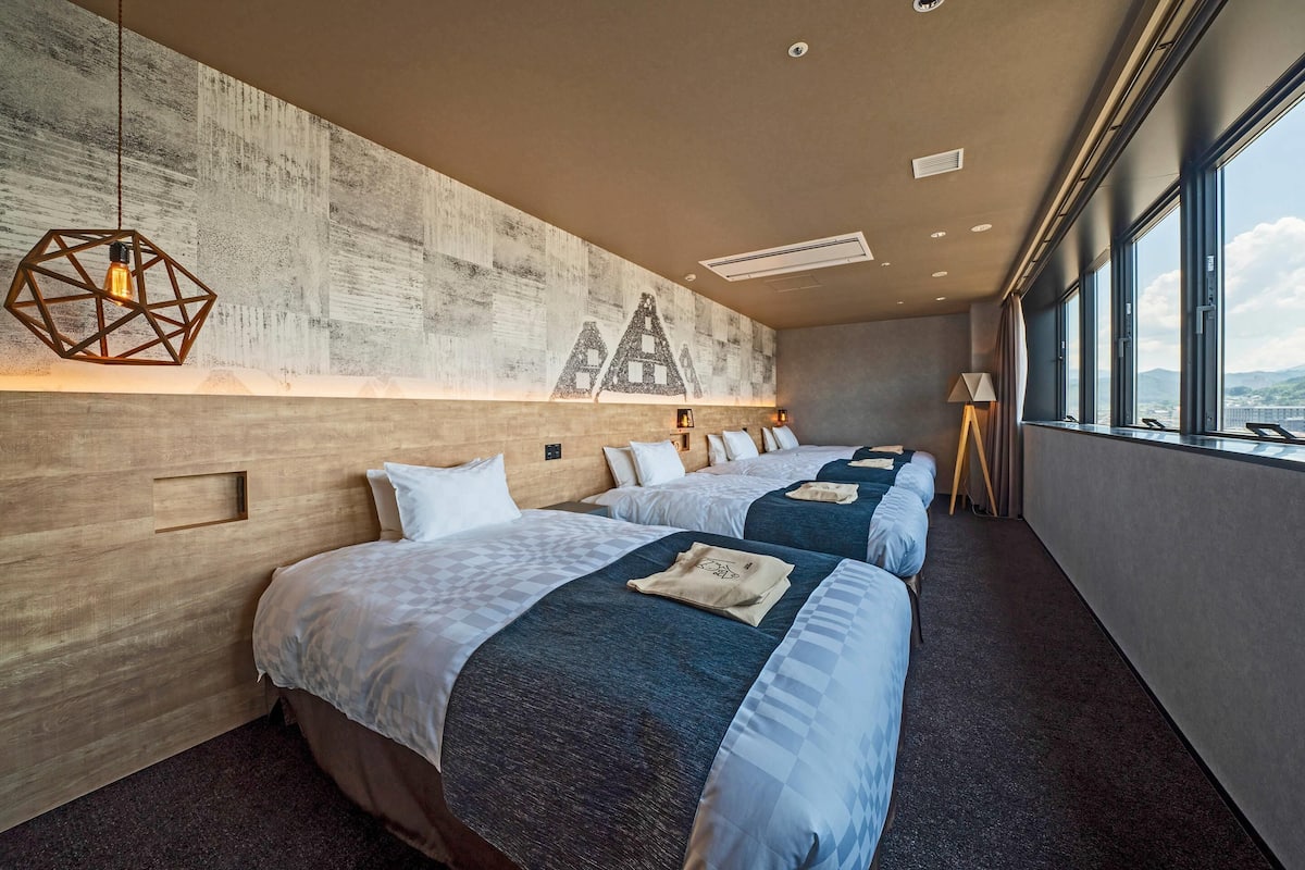 A hotel room with three beds, a concrete wall with geometric patterns, and large windows offering a view.