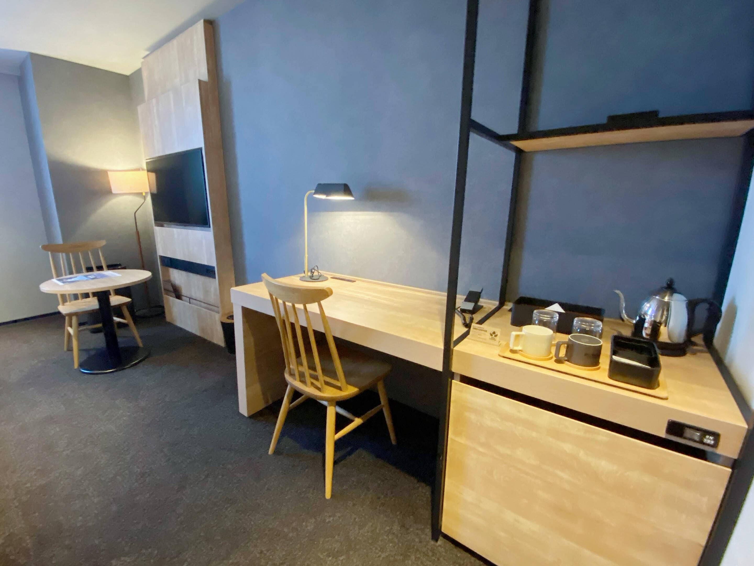 A compact hotel room with a single bed, a small desk with a chair, a bedside table with a lamp, and a kitchenette area.