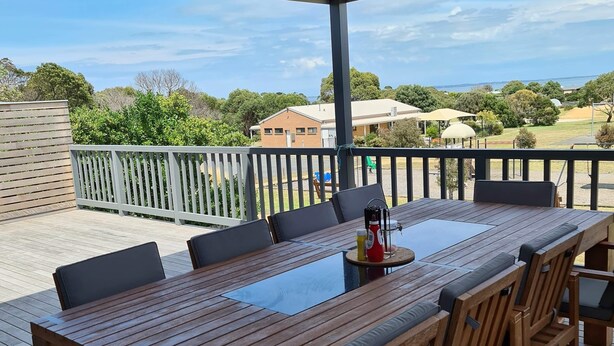 A wooden deck with a table and chairs, overlooking a residential area with houses and trees. - 4 bedrooms, large deck, park views (Coronet Bay)