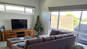 A living room with a sectional sofa, a coffee table, a TV on a wooden stand, a Christmas tree, and a large window with a view of the outdoors. - 4 bedrooms, large deck, park views (Coronet Bay)