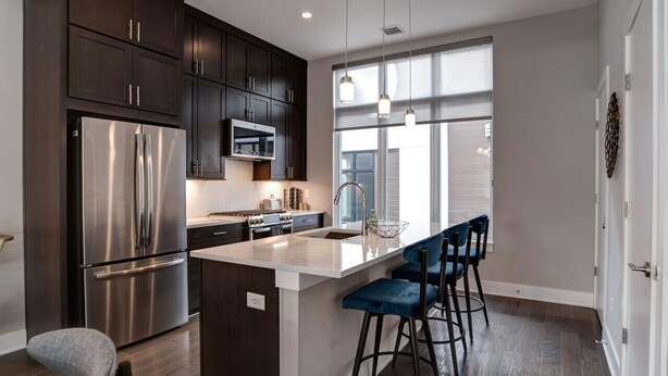 A modern kitchen with dark cabinets, stainless steel appliances, a large island with bar stools, and a window providing natural light. - South Slope Skyhaus with Rooftop Terrace (Asheville)