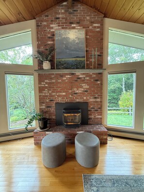 A room with a brick fireplace, large windows, and a view of greenery. - Serene mountain escape with beautiful views and a hot tub (Jefferson)