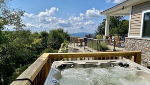 Un jacuzzi con terraza de madera, rodeado de vegetación y un muro de piedra, con vista a montañas al fondo.