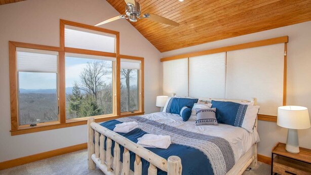 Un dormitorio con techo de madera, cama con ropa de cama azul, dos ventanas y un ventilador de techo.