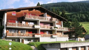 Un immeuble à plusieurs étages doté de balcons, d’un espace de stationnement et ayant une chaîne de montagnes en arrière-plan.