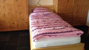 Un lit simple avec une couverture à motifs, une armoire en bois et une petite lampe de bureau.