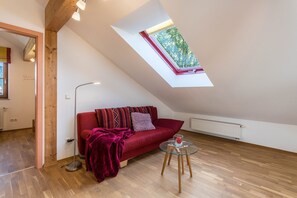 Une pièce avec un plafond en pente, un canapé rouge, une table en verre et une fenêtre donnant sur des arbres.