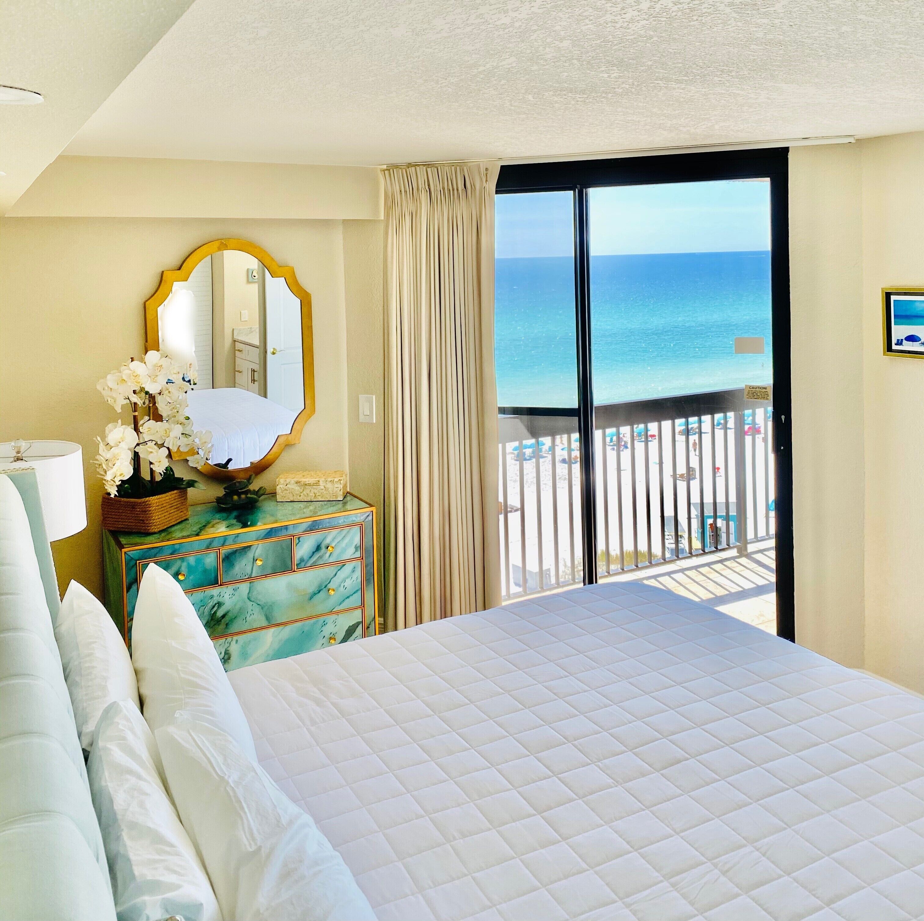 A hotel room with a bed, a dresser, a mirror, and a view of the beach.