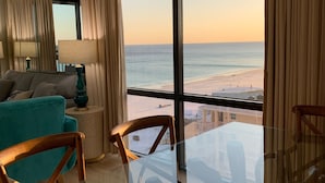 A modern living room with a glass table, teal sofa, and wooden chairs. A large window offers a view of the beach.