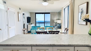 A spacious living area with a large window overlooking the ocean, a ceiling fan, and a dining area with a marble countertop.