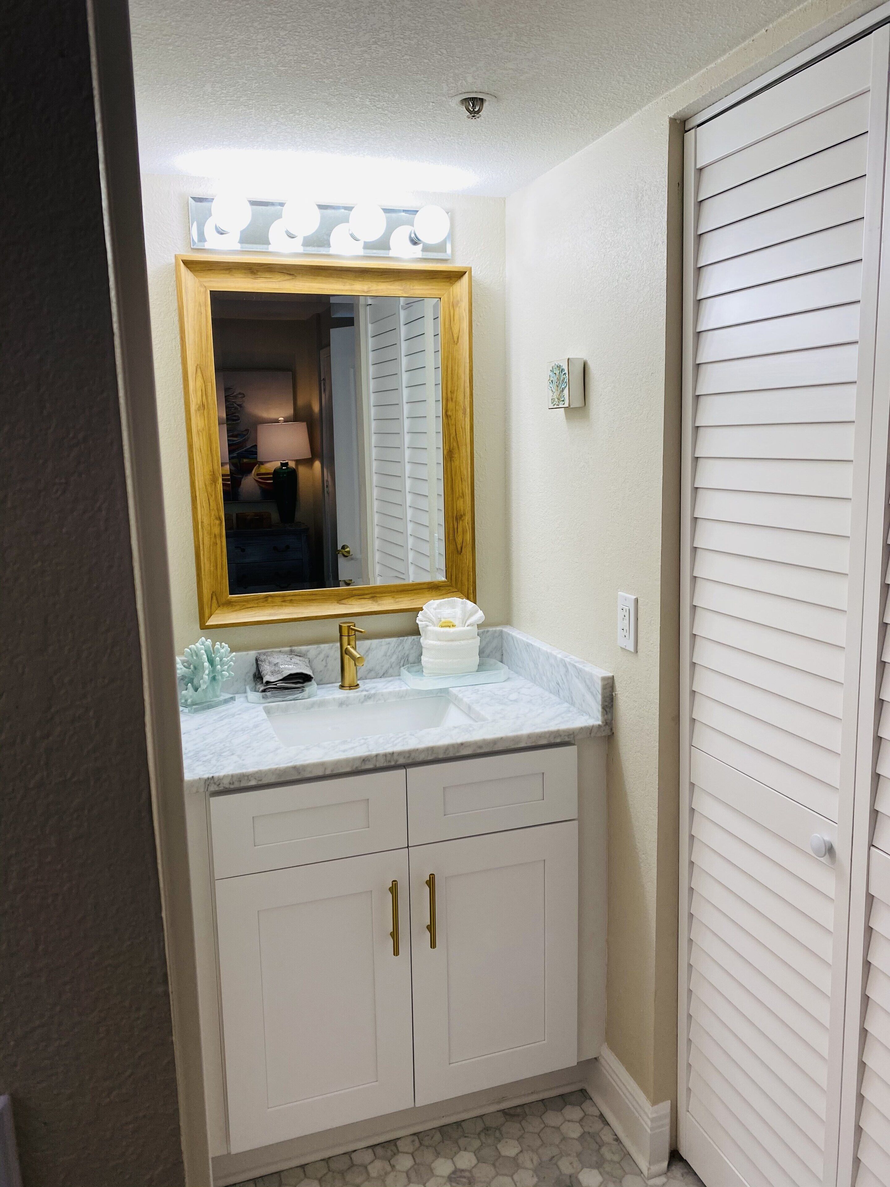A bathroom with a vanity, a mirror with lights, a sink, and a door with blinds.