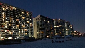 A multi-story hotel with numerous illuminated windows at night, a sandy beach in the foreground, and a clear sky.