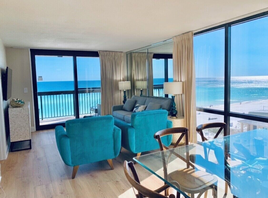 A modern hotel room with a balcony overlooking the ocean, a glass dining table, and a comfortable seating area.