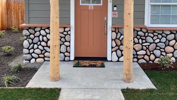 A house with a stone foundation, a brown front door, and a porch with wooden pillars. - Riverside Cabin 4 - (Brand New Construction!) (Grants Pass)