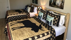 A double room with two beds, each with a checkered bedspread featuring a deer silhouette. There is a nightstand with a lamp, a painting on the wall, and an air conditioning unit above the beds. - Riverside Cabin 4 - (Brand New Construction!) (Grants Pass)