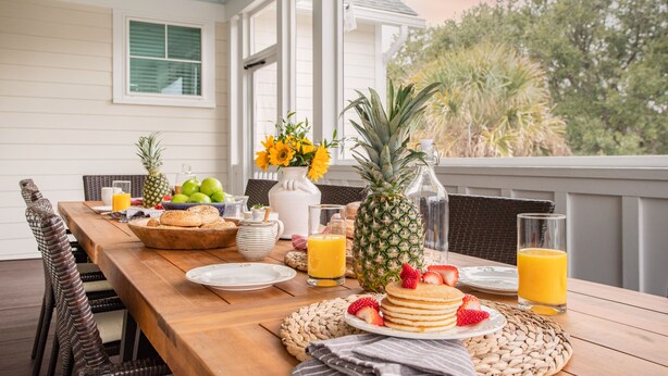 Una mesa de desayuno con tortitas, fruta y zumo de naranja en una terraza de madera con barandas blancas y vista a árboles.