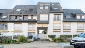 Un immeuble résidentiel à plusieurs étages, doté d’une entrée centrale, de balcons et d’un espace de stationnement où se trouve une voiture blanche.