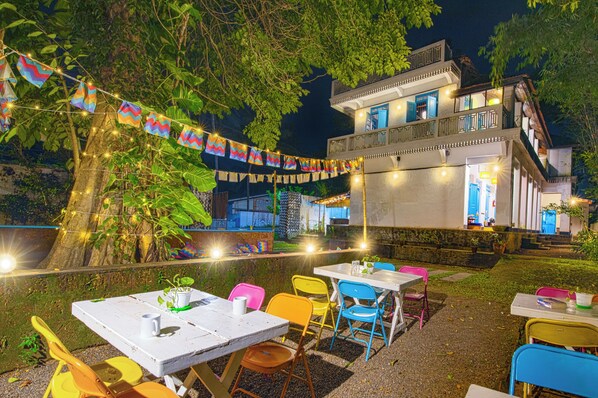 An outdoor dining area with tables and chairs, string lights, and a building in the background.