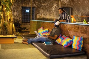 A person using a laptop on a sofa with colorful pillows, a laptop on a table, and a bar area with a drink. - goSTOPS Lite Alleppey (Ambalapuzha)