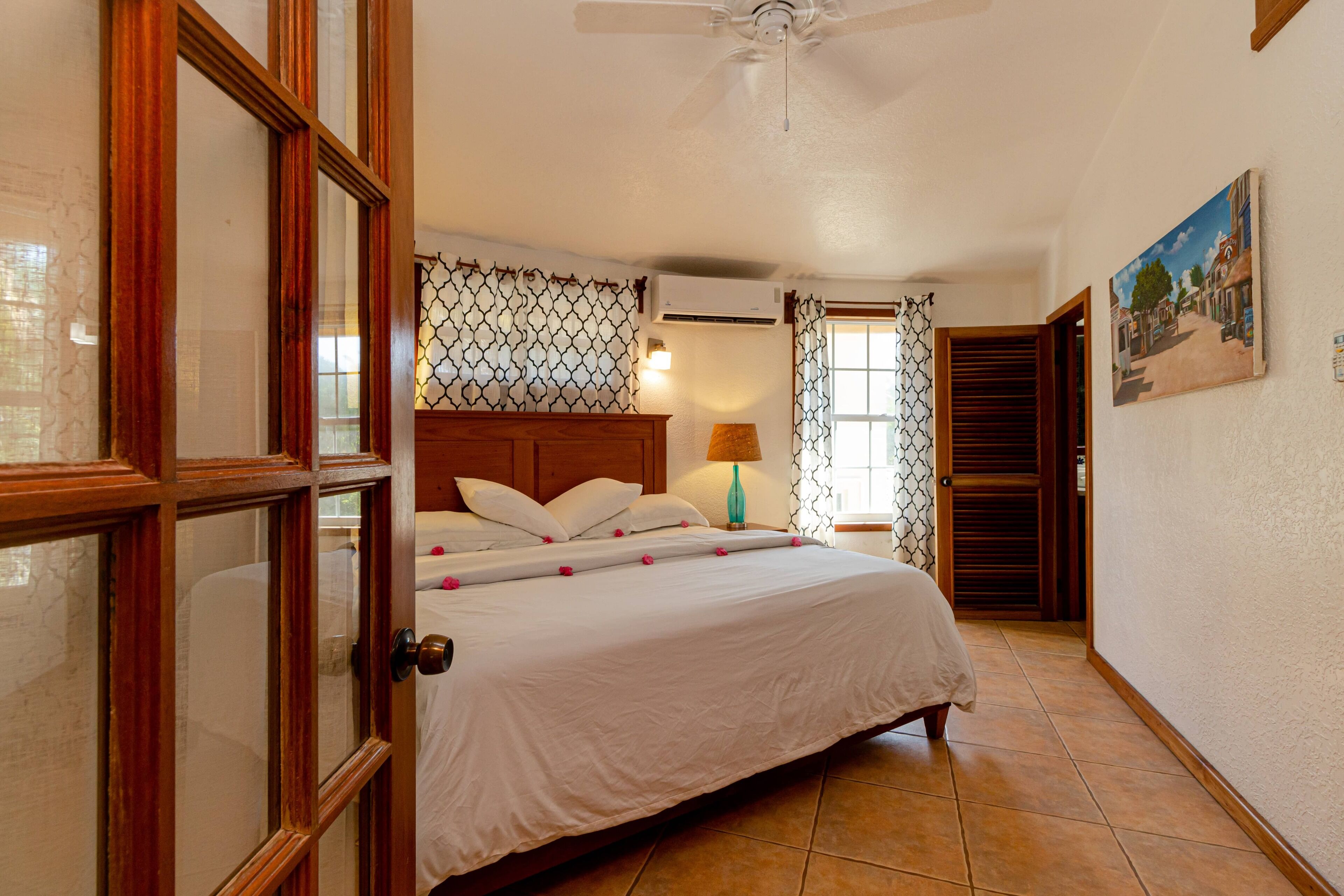 Un dormitorio con cama, ventilador de techo, ventana con cortinas y un cuadro en la pared.