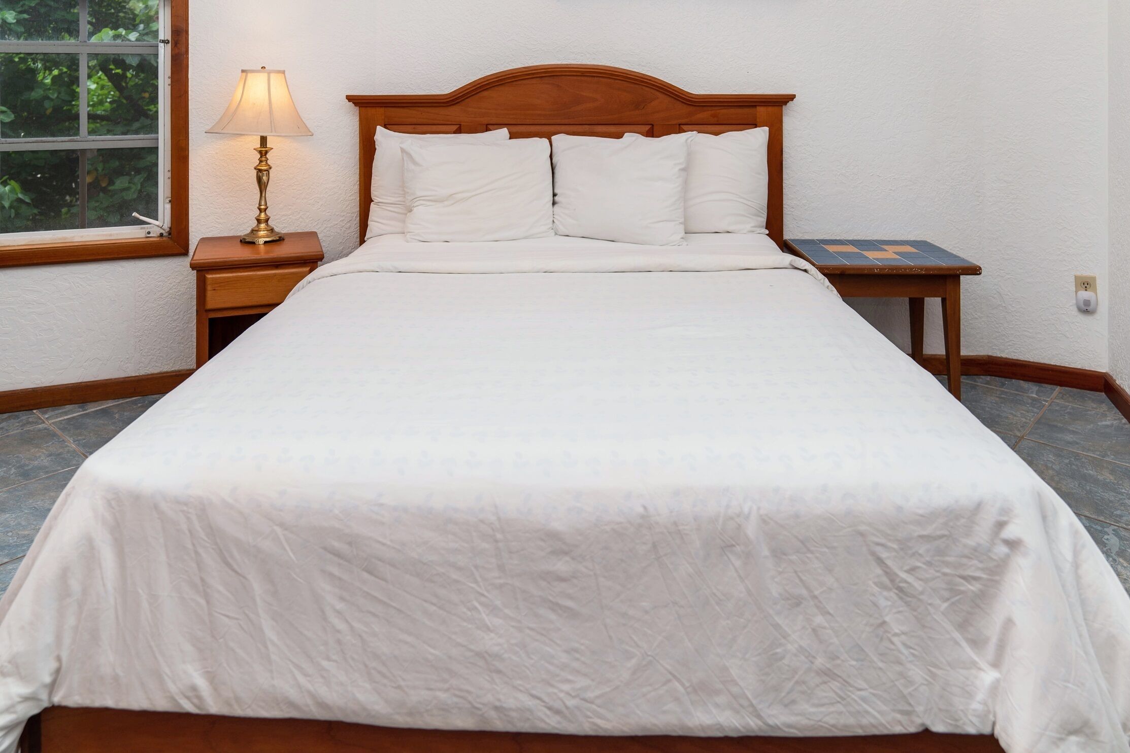 Een keurig opgemaakt bed met witte lakens, een houten hoofdbord, een bedlampje en een klein tafeltje met een blauw-oranje geruit tafelkleed.