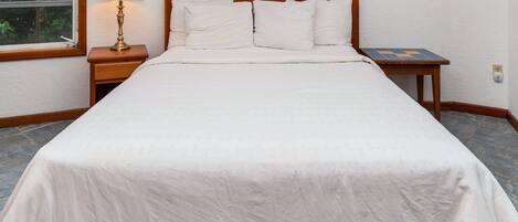 Een keurig opgemaakt bed met witte lakens, een houten hoofdbord, een bedlampje en een klein tafeltje met een blauw-oranje geruit tafelkleed.