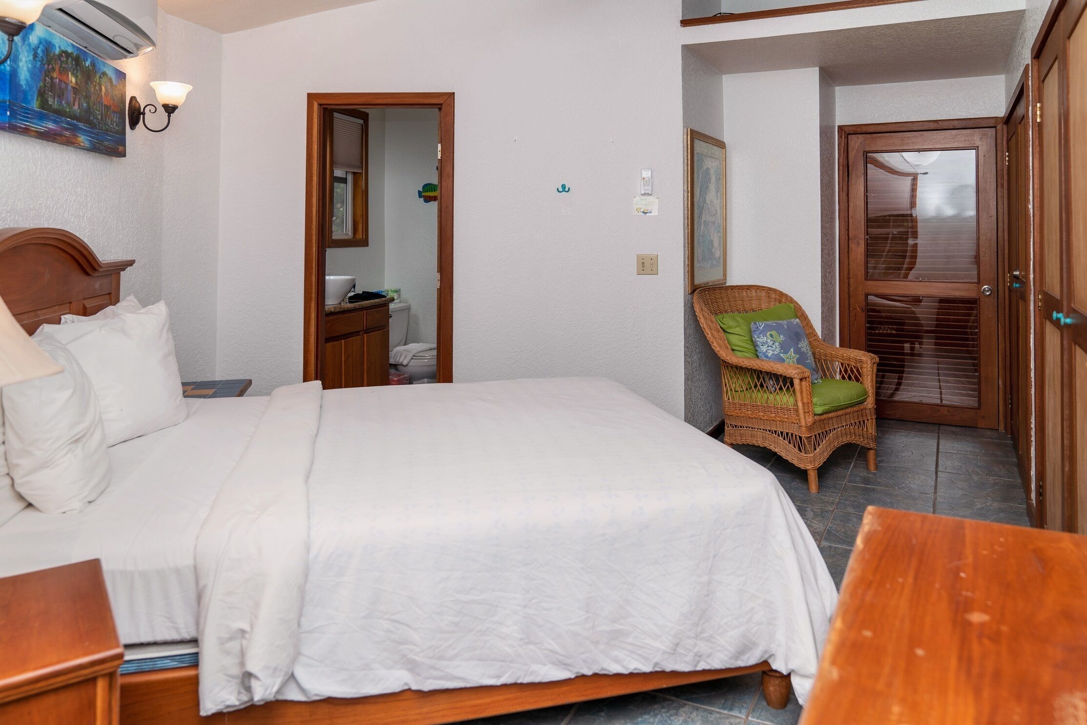 A neatly made bed with white linens, a wooden nightstand, a wicker chair with a cushion, a small table, and a door leading to a bathroom.