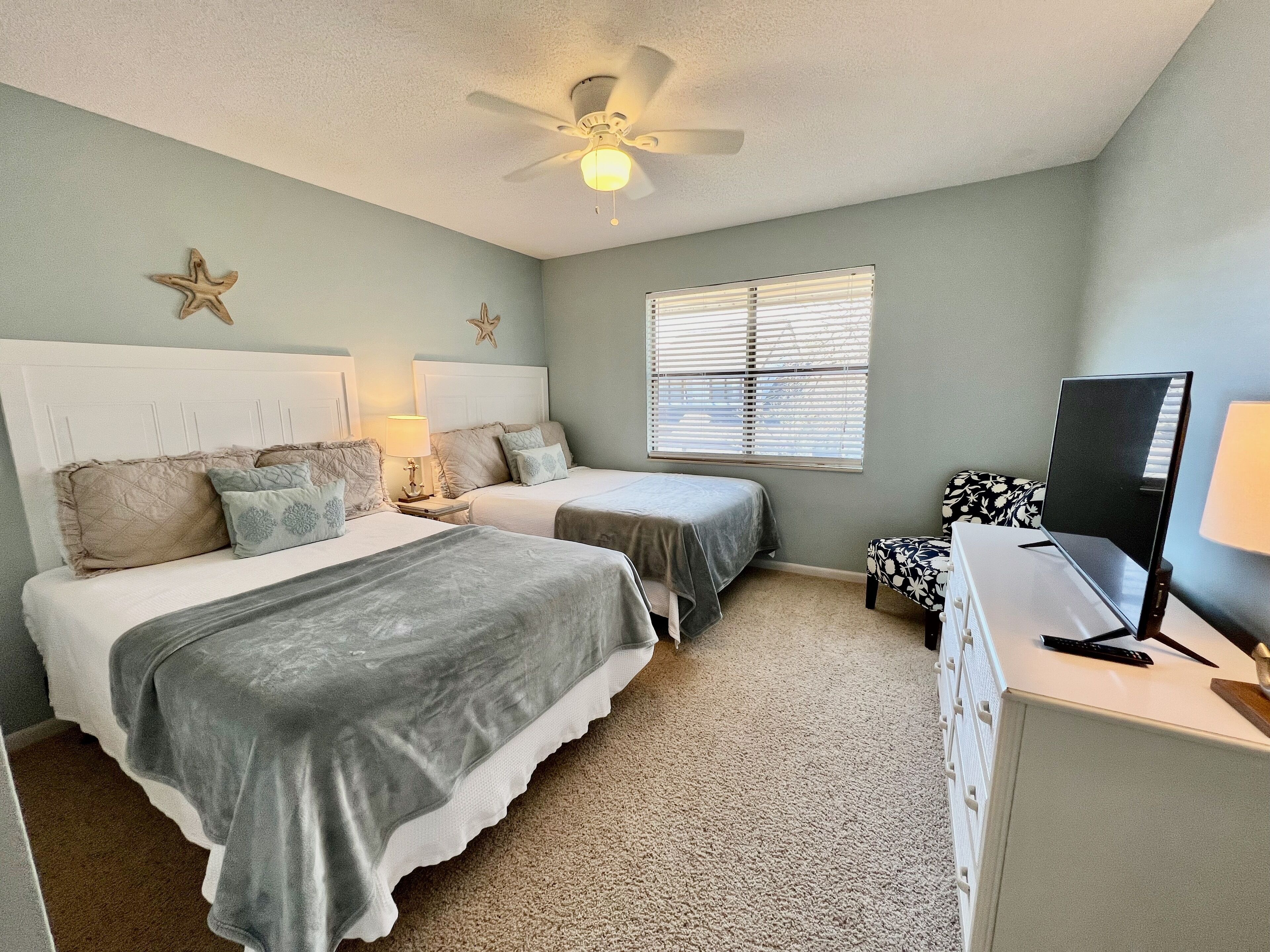 A bedroom with two beds, a ceiling fan, a window with blinds, a desk with a computer, and a starfish decoration on the wall.