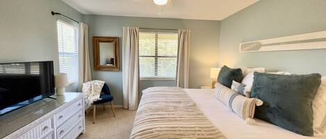 Quarto com cama, televisão, ventoinha no teto, cômoda e janela com cortinas.