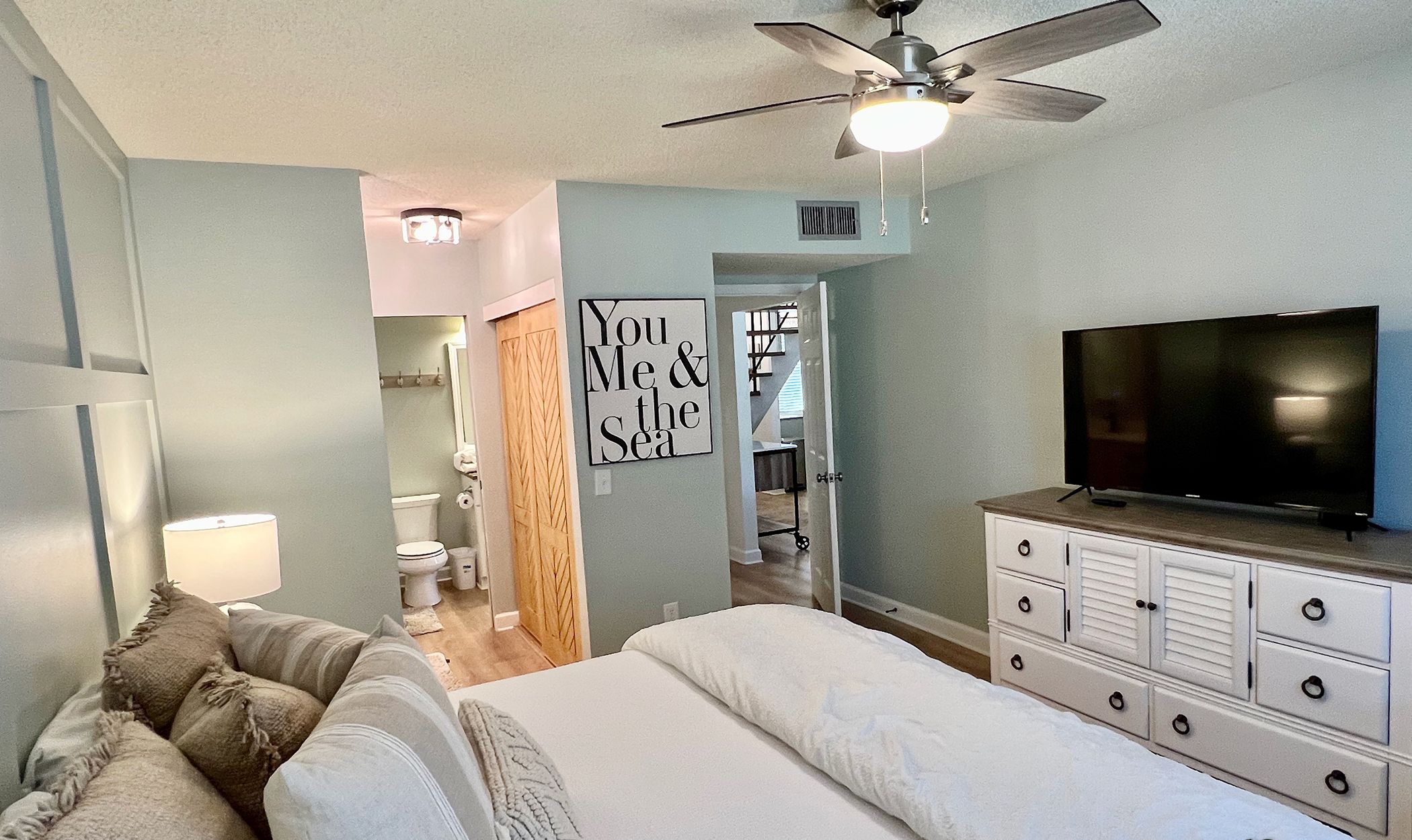 A bedroom with a bed, a TV, a ceiling fan, and a sign that reads "You Me & the Sea".