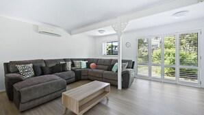 Un salon moderne avec un canapé d’angle, une table basse en bois et de grandes fenêtres équipées de stores.