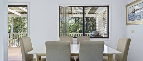 Un espace repas avec une table blanche et six chaises, de grandes fenêtres donnant sur la forêt et une terrasse en bois à l’extérieur.