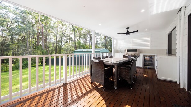 Une terrasse extérieure couverte avec une plateforme en bois, une table à manger avec des chaises, un ventilateur de plafond et une vue sur une pelouse verdoyante et des arbres.