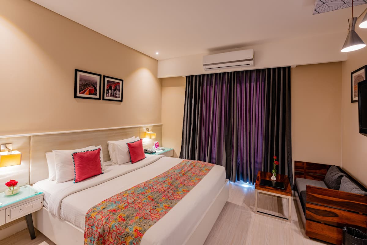 A hotel room with a large bed, a bedside table with a lamp, a small table with a vase of flowers, and a sofa.