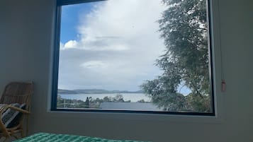 A room with a large window offering a view of a body of water, a chair, and a bed with a green and white patterned bedspread.