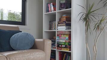 A room with a beige leather sofa, a blue pillow, a bookshelf with various board games, and a potted plant.