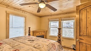Un dormitorio con techo de madera, ventilador de techo, cama con colcha de estilo vintage, chimenea y ventanas grandes.