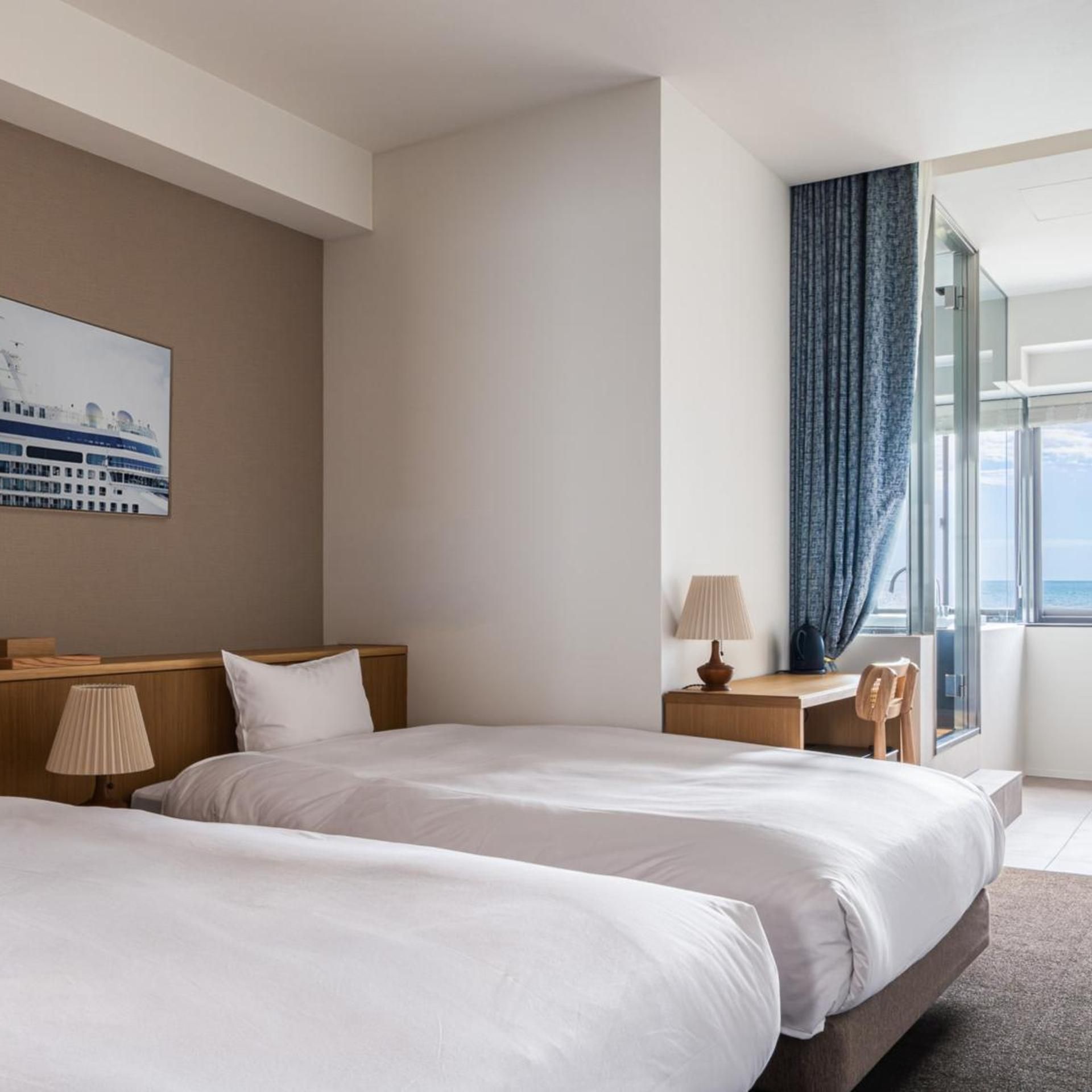 A hotel room with two beds, a desk, a chair, a lamp, a window with curtains, and a painting on the wall.