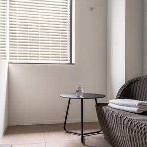 Une pièce avec une fenêtre, une table ronde surmontée d’un plateau en verre et une chaise en osier.