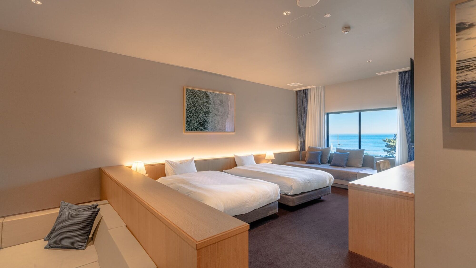 Habitación de hotel con dos camas, una zona de estar, un ventanal con vistas y un cuadro en la pared.