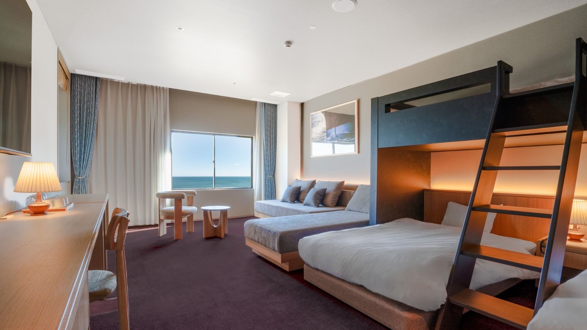 Habitación de hotel con literas, un escritorio con lámpara, una mesita pequeña, dos sillas, un sofá y un ventanal con vistas.