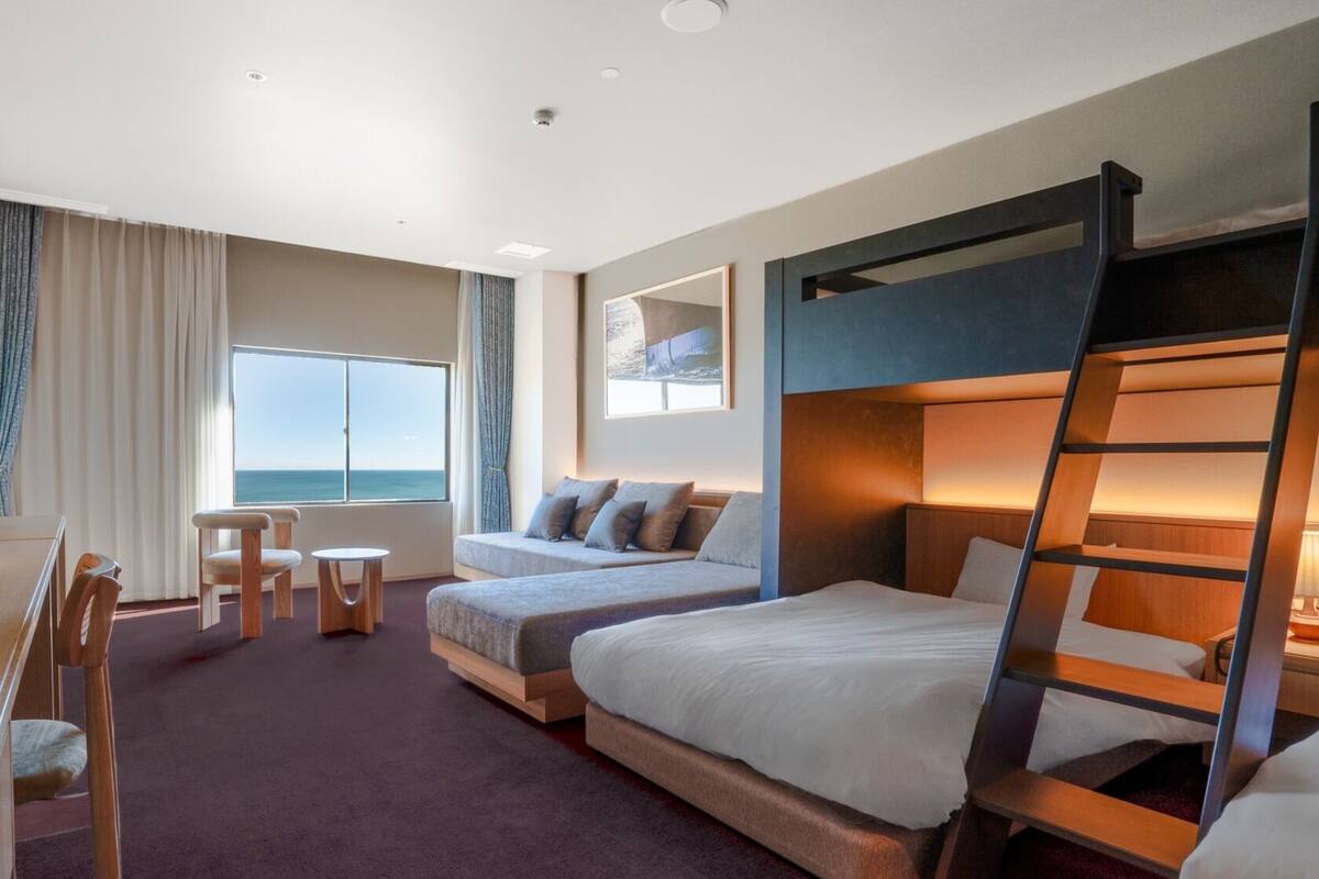 Een hotelkamer met een stapelbed, een bureau met lamp, een bijzettafeltje, twee stoelen, een bank en een groot raam met uitzicht.