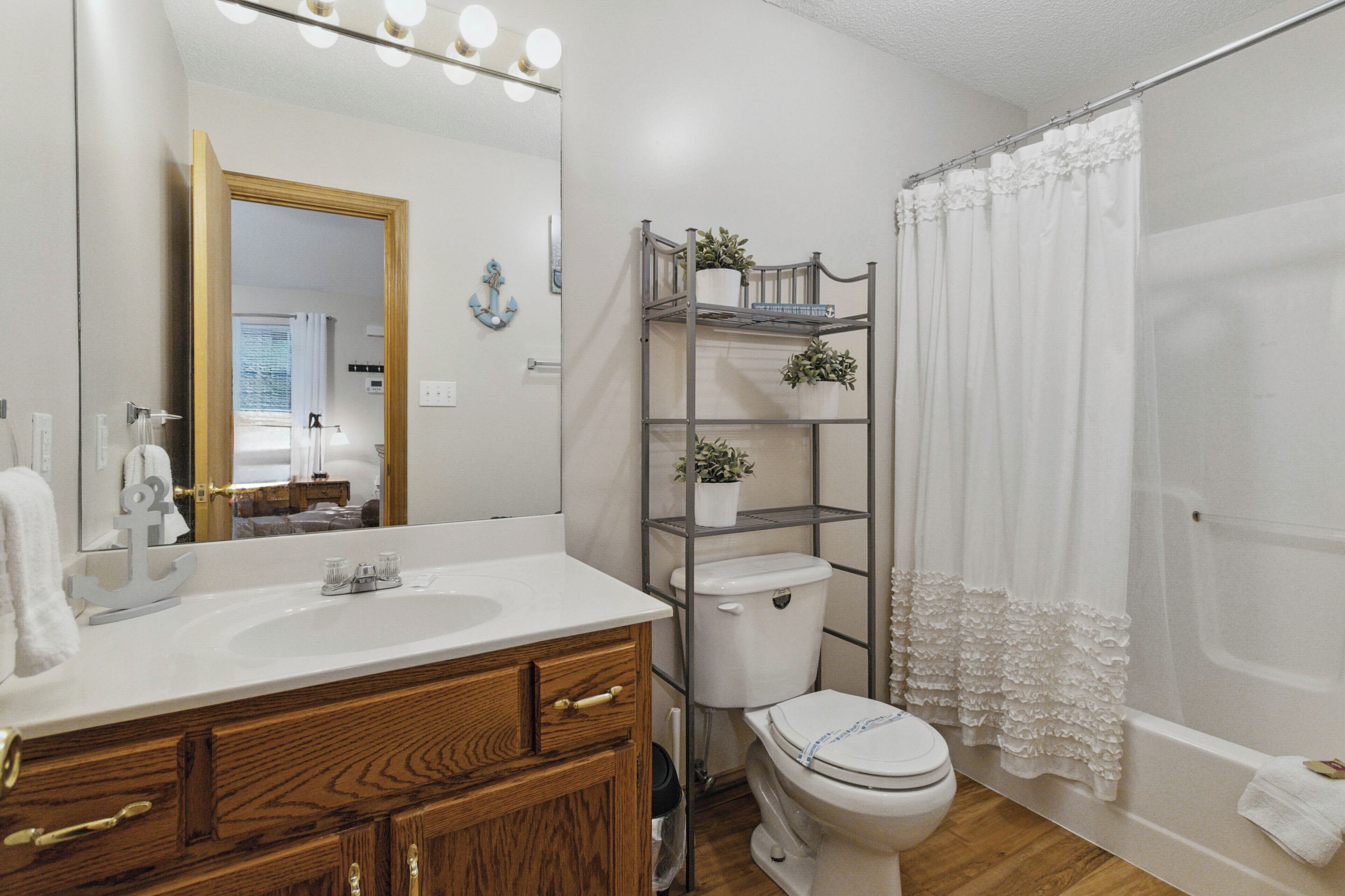 A bathroom with a wooden vanity, a white sink, a toilet, and a shower with a white curtain. There are plants on a metal shelf above the toilet.