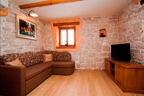 A cozy room with a stone wall, a wooden ceiling, a brown sofa with patterned cushions, a TV on a wooden stand, and a window with a plant. - Authentic Villa Ulika with a pool and a jacuzzi (Visnjan)