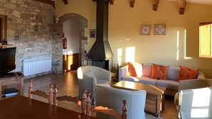 Un salon chaleureux avec une cheminée en pierre, des poutres en bois, un mur en pierre, une télévision et un canapé agrémenté de coussins orange.