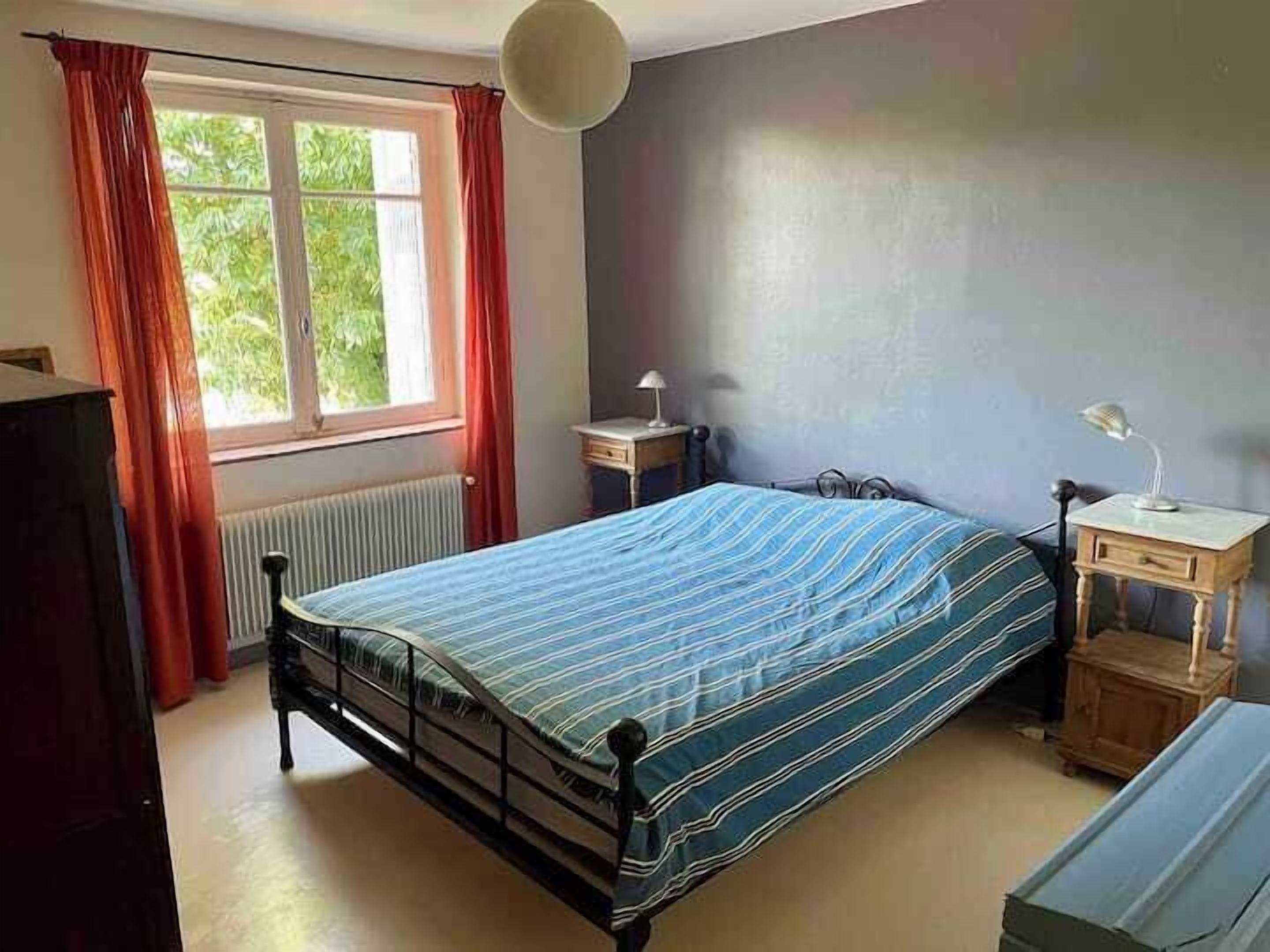 A bedroom with a bed, a window with curtains, a radiator, a bedside table with a lamp, and a dresser.