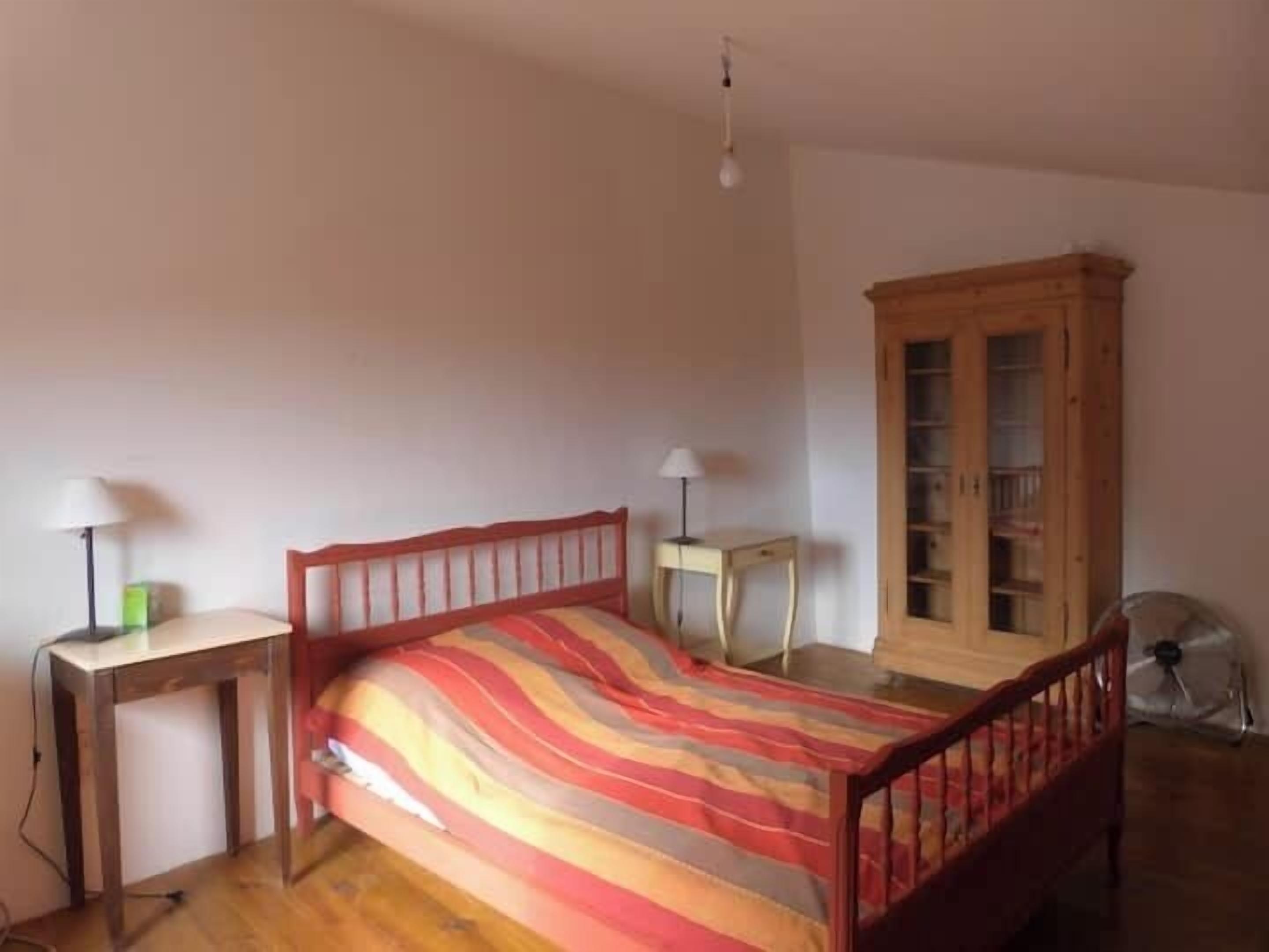 A bedroom with a wooden bed, a bedside table with a lamp, a cabinet, and a floor fan.